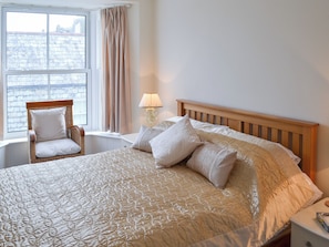 A bedroom with a large bed, a wooden headboard, a wicker chair with cushions, a bedside table, and a window with curtains.