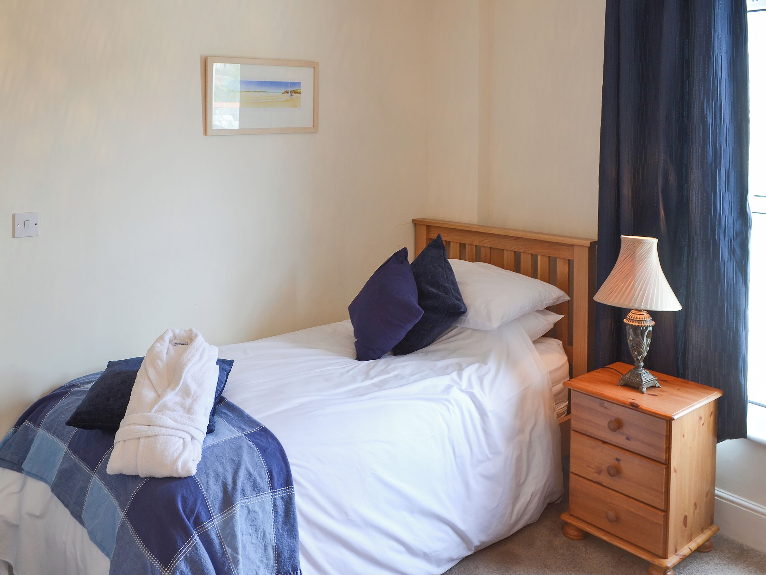 A neatly made bed with a blue and white checkered blanket, a wooden nightstand with a lamp, and a framed picture on the wall.