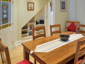 A dining area with a wooden table, chairs, and a bowl on a placemat. A bookshelf with books and a framed picture are visible in the background.