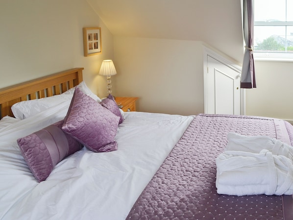 A neatly made bed with white and purple bedding, a bedside lamp, and a window with curtains. - 3 bedroom accommodation in Padstow (Padstow)