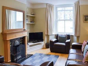 A living room with a fireplace, television, leather sofa, and a large window with curtains. - 3 bedroom accommodation in Padstow (Padstow)