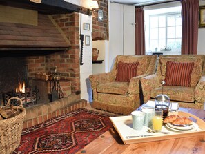A cozy living room with a fireplace, two patterned armchairs, a wooden coffee table with breakfast items, and a red patterned rug. - Tranquillity Cottage (Winfrith Newburgh)
