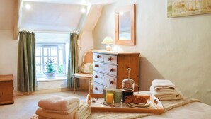 Une chambre à coucher avec un lit, une commode en bois, une fenêtre avec des rideaux, une table de chevet avec une lampe, et un plateau avec des serviettes et des articles pour le petit-déjeuner.