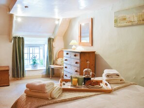 A bedroom with a bed, a wooden dresser, a window with curtains, a bedside table with a lamp, and a tray with towels and breakfast items.