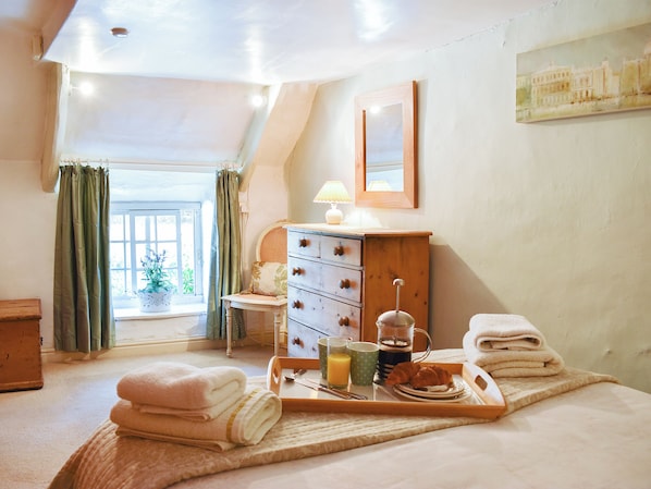 A bedroom with a bed, a wooden dresser, a window with curtains, a bedside table with a lamp, and a tray with towels and breakfast items.
