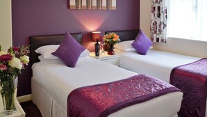A hotel room with two beds, purple bedding, a nightstand with a lamp and a vase of flowers, and a painting on the wall. - 3 bedroom accommodation in Liskeard (Liskeard)