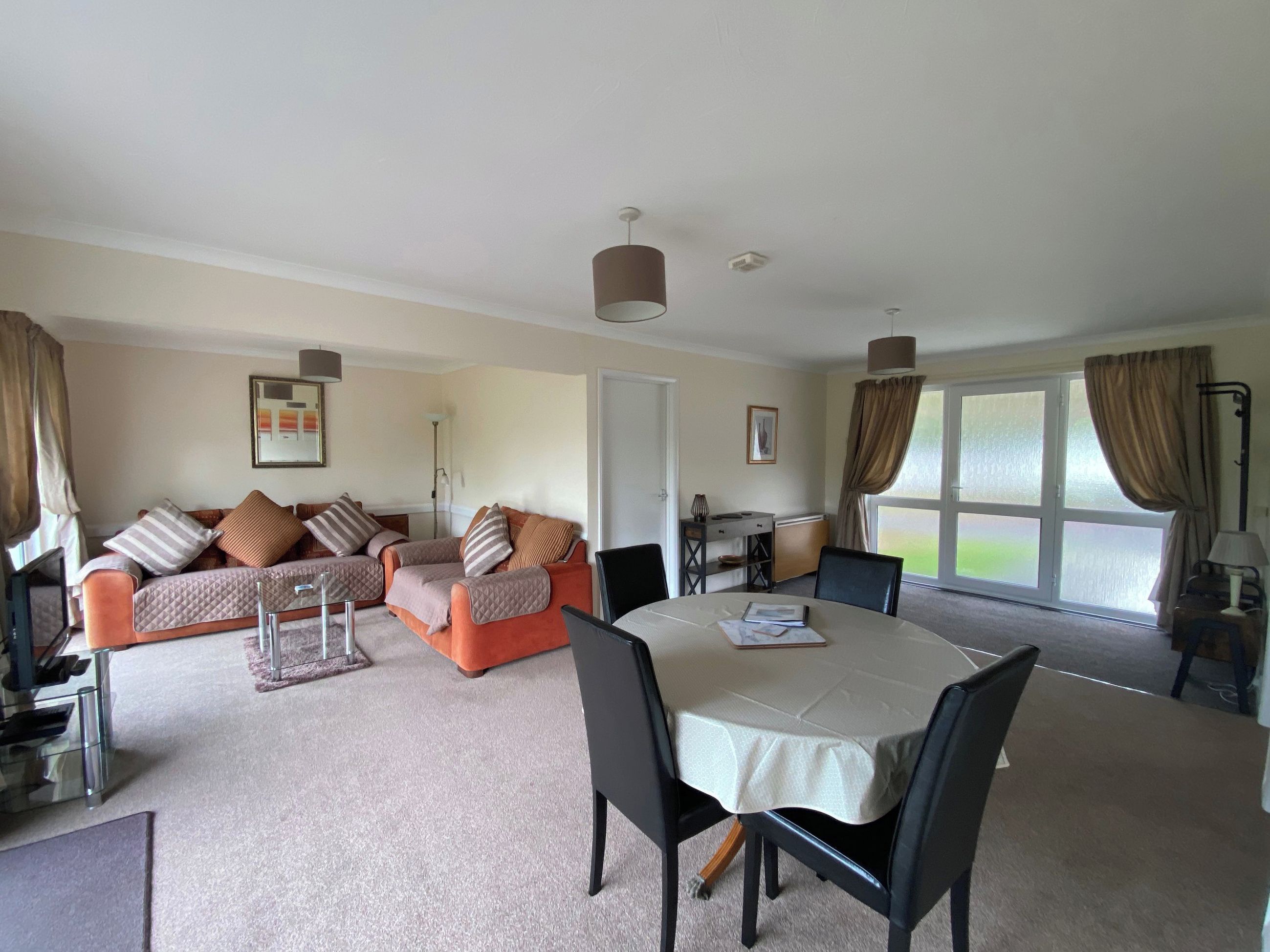 A spacious living area with a sofa set, a dining table with chairs, a lamp, and a large window with curtains.