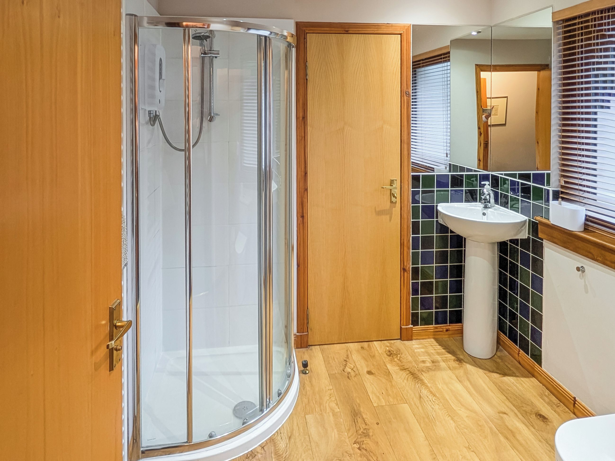 A modern bathroom with a glass-enclosed shower, a wooden door, a sink with a mirror, and tiled walls.