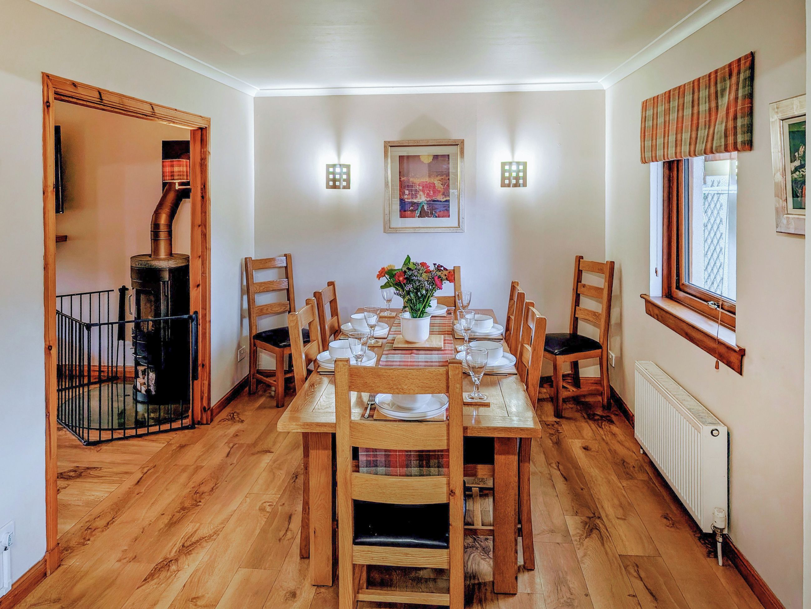 A dining room with a wooden table set for six, surrounded by wooden chairs. There is a fireplace on the left, a radiator on the right, and a window with a plaid valance.