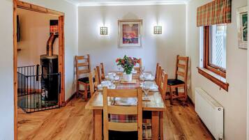 A dining room with a wooden table set for six, surrounded by wooden chairs. There is a fireplace on the left, a radiator on the right, and a window with a plaid valance.