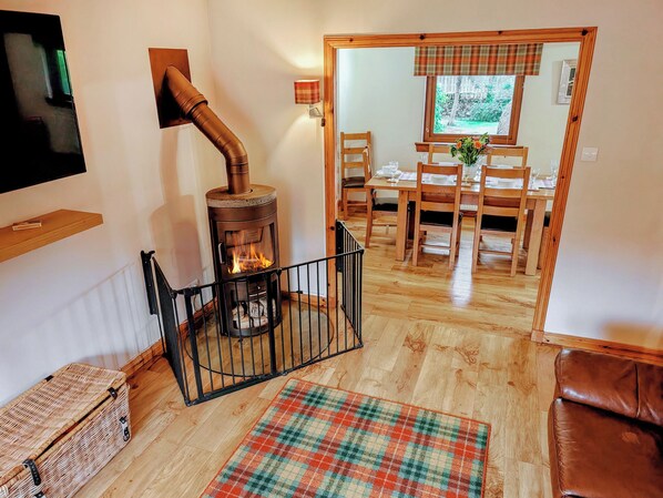 A cozy living room with a wood-burning stove, a dining area with wooden furniture, and a view of the outdoors through a window. - 3 bedroom accommodation in Carrbridge (Carrbridge)