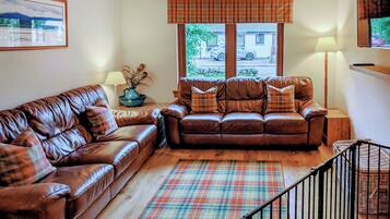 A living room with a brown leather sofa, a matching armchair, a plaid rug, and a window with a view of the outside.