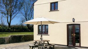 A patio with a table and chairs, a white umbrella, and a building with a door and windows. - 4 bedroom accommodation in Bradworthy, near Hartland (Bradworthy, near Hartland)