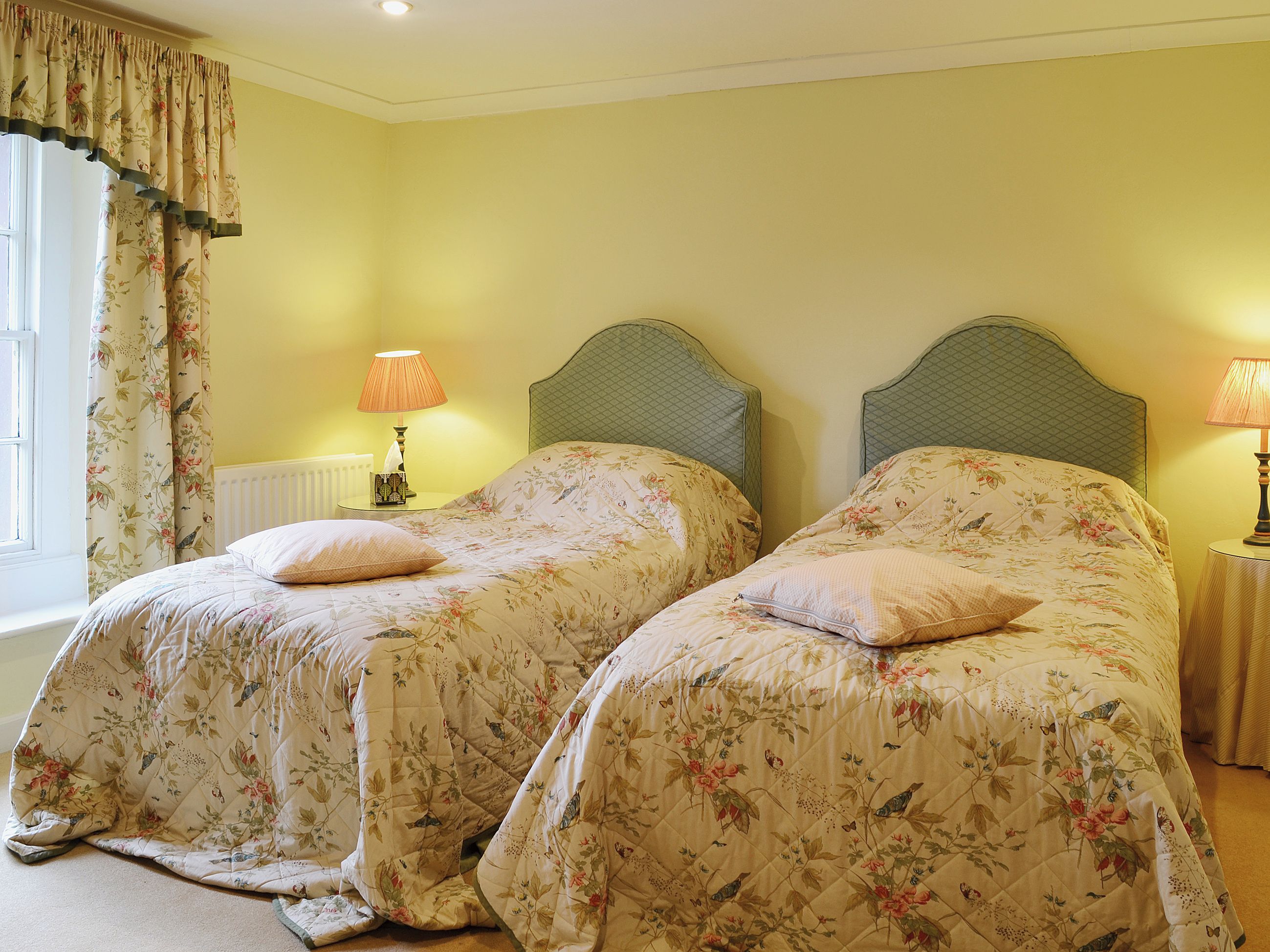 A room with two beds, each with floral bedding and a matching headboard. There are bedside lamps and a window with curtains.