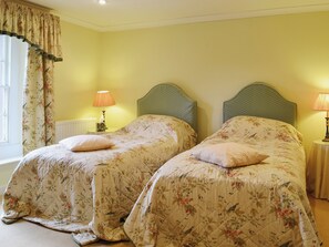 Una habitación con dos camas, cada una con ropa de cama floral y un cabecero a juego. Hay lámparas de noche y una ventana con cortinas.