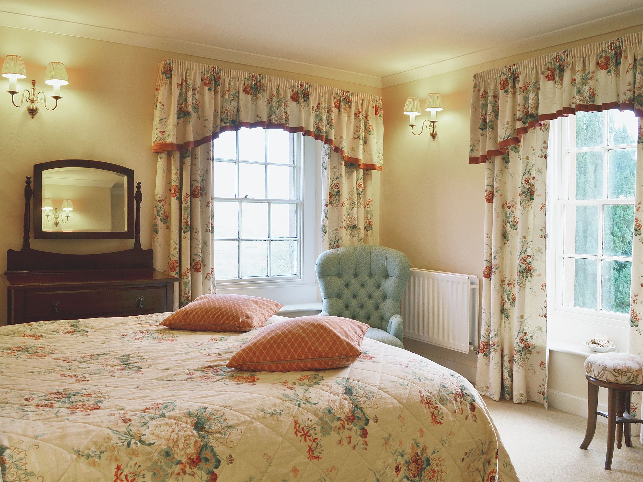Ein Schlafzimmer mit einem großen Bett, einem Sessel, einem Schminktisch, einem Spiegel und zwei mit Vorhängen versehenen Fenstern.