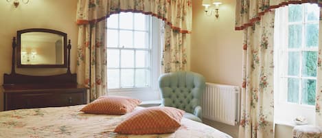A bedroom with a large bed, a chair, a dresser, a mirror, and two windows with curtains.