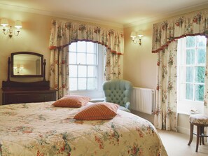 A bedroom with a large bed, a chair, a dresser, a mirror, and two windows with curtains.