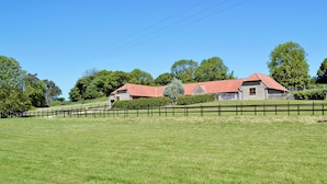 A stone farmhouse with a red roof, surrounded by a wooden fence and lush green fields.