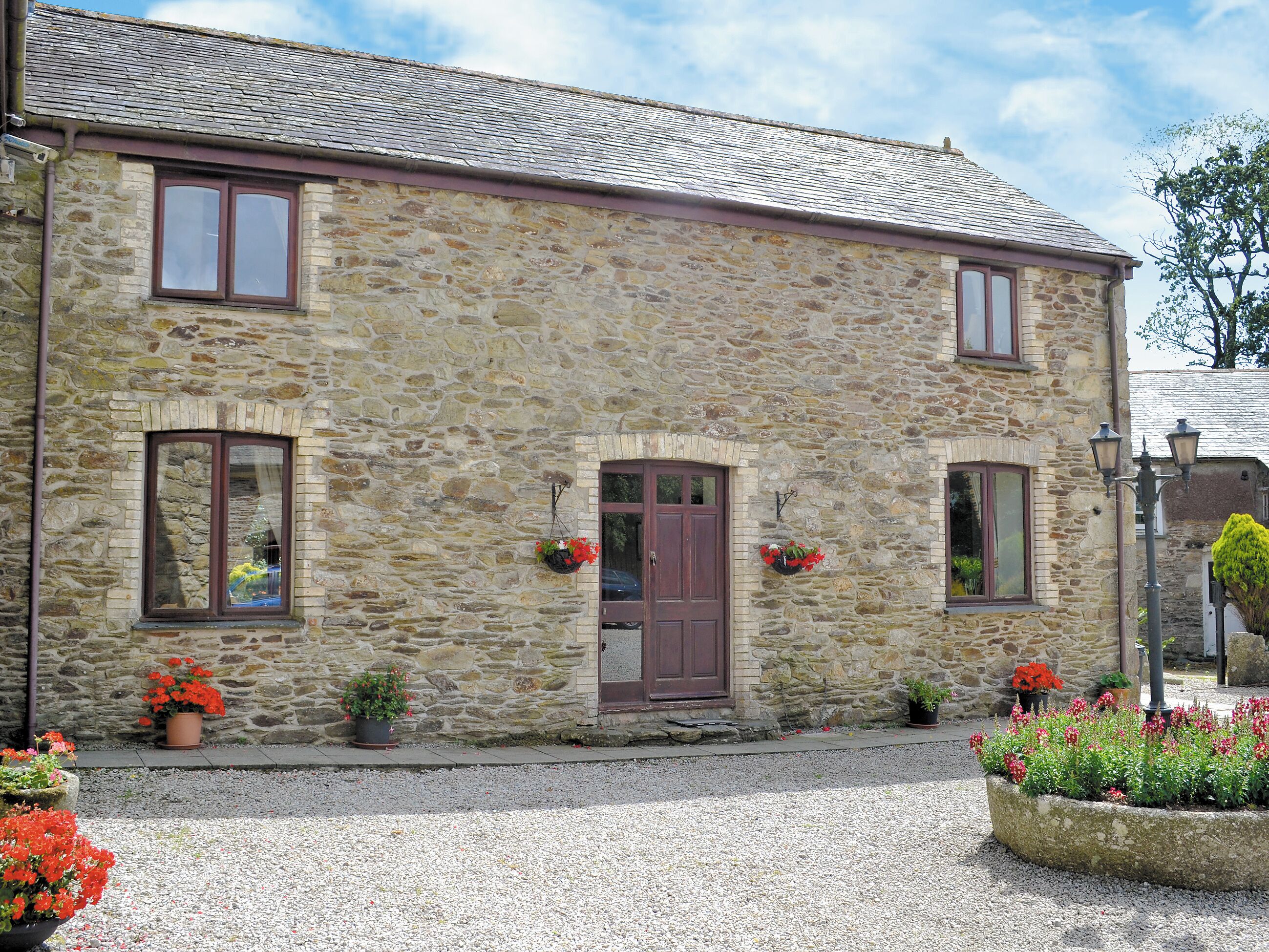 A stone building with a traditional design, featuring a central entrance with a wooden door, flanked by windows with brown frames. There are flower pots with red flowers placed on the ground and hanging on the wall.