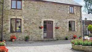 A stone building with a traditional design, featuring a central entrance with a wooden door, flanked by windows with brown frames. There are flower pots with red flowers placed on the ground and hanging on the wall. - 3 bedroom accommodation in Mevagissey, near St. Austell (Mevagissey, near St. Austell)