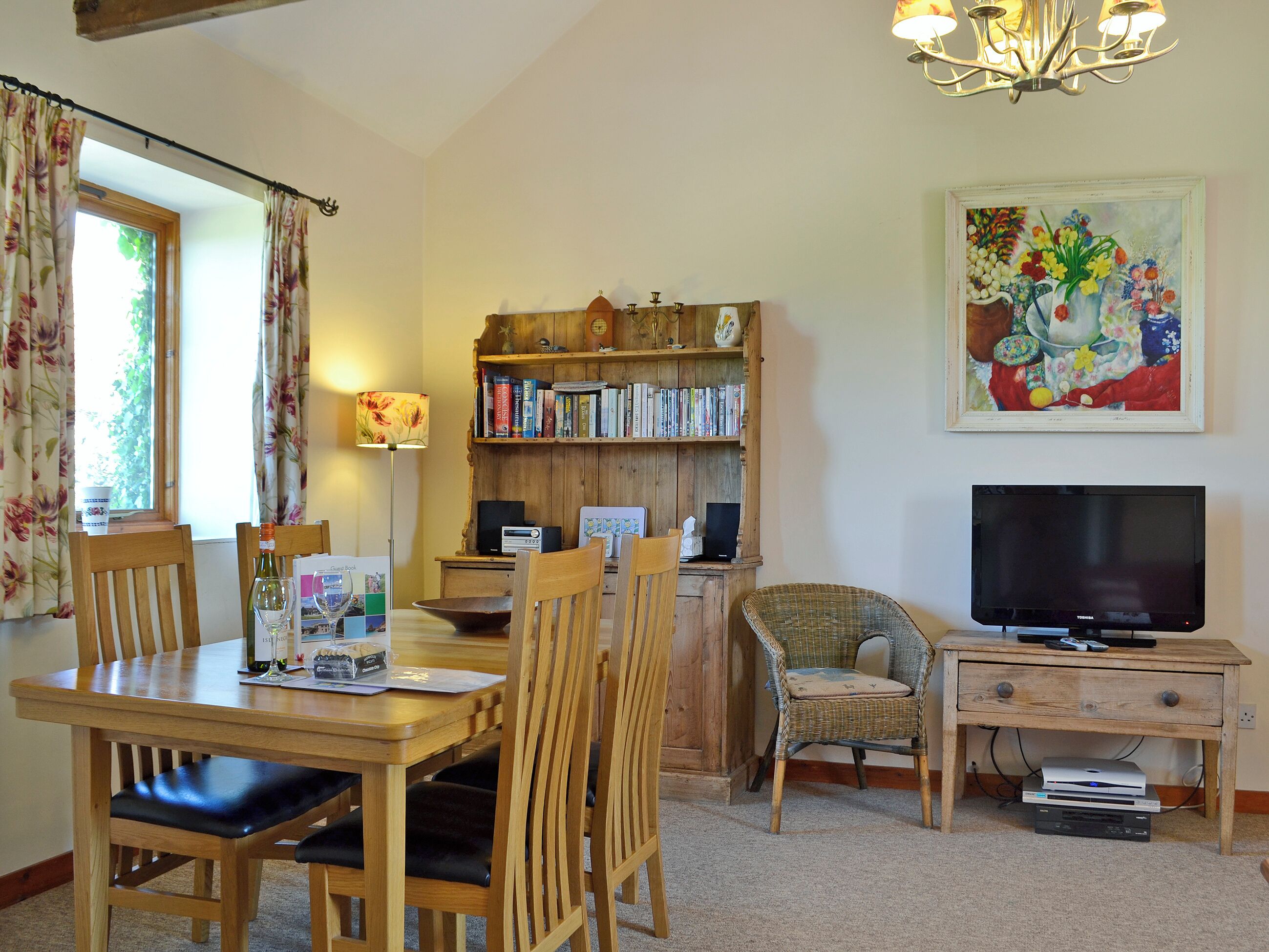 A cozy living room with a wooden dining table, comfortable chairs, a bookshelf, a television, and a painting on the wall.
