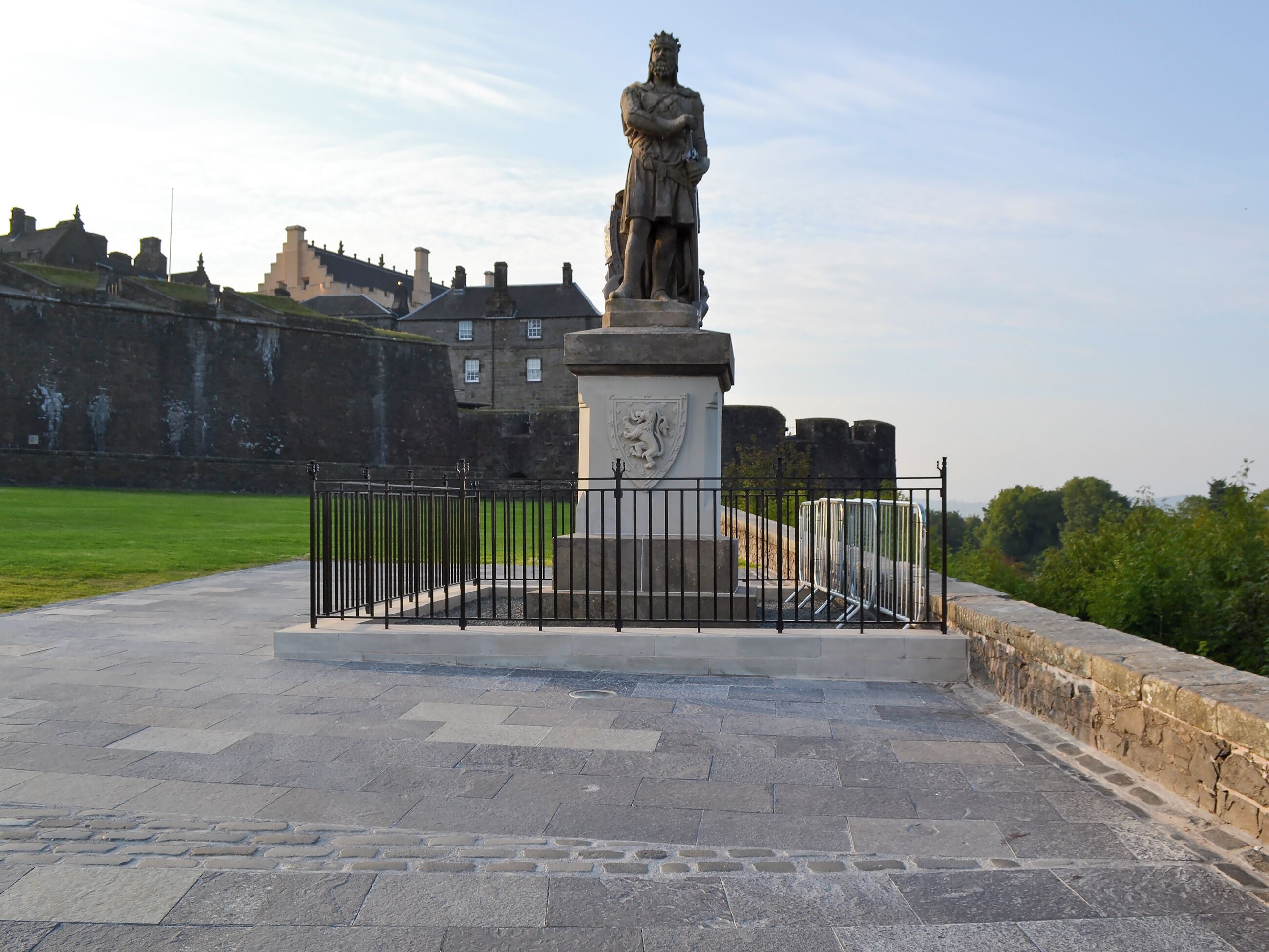 Une statue sur un piédestal, protégée par une barrière, se dressant devant un décor composé d’immeubles et d’un ciel dégagé.