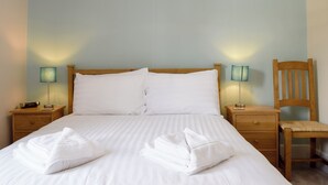 Una cama bien tendida con sábanas blancas y dos almohadas, una mesita de noche de madera con una lámpara, y una silla de madera.