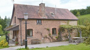 A stone house with a sloped roof, a chimney, and a small porch with a bench. - 4 bedroom accommodation in Lea, near Ross-on-Wye (Lea, near Ross-on-Wye)