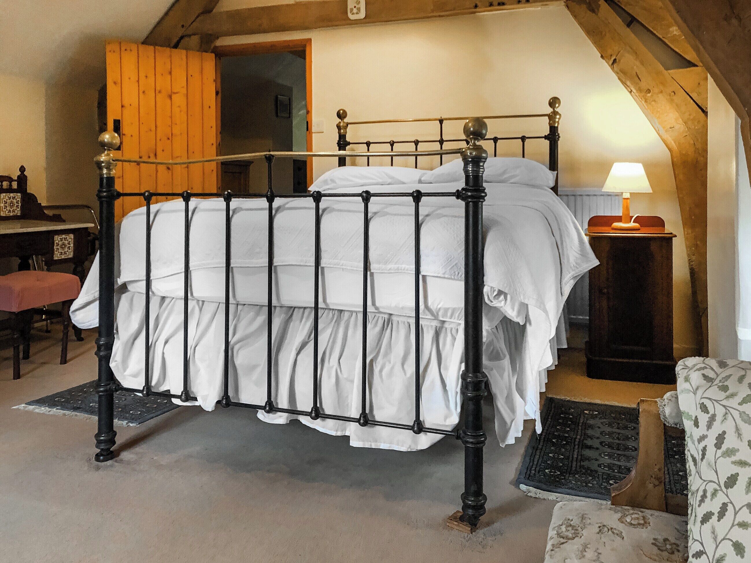 Une chambre à coucher avec un lit en métal, du linge de lit blanc, une porte en bois, un bureau avec une chaise et une lampe.