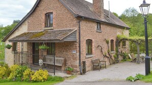 A stone house with a sloped roof, a chimney, and a small porch with a bench. - 4 bedroom accommodation in Lea, near Ross-on-Wye (Lea, near Ross-on-Wye)