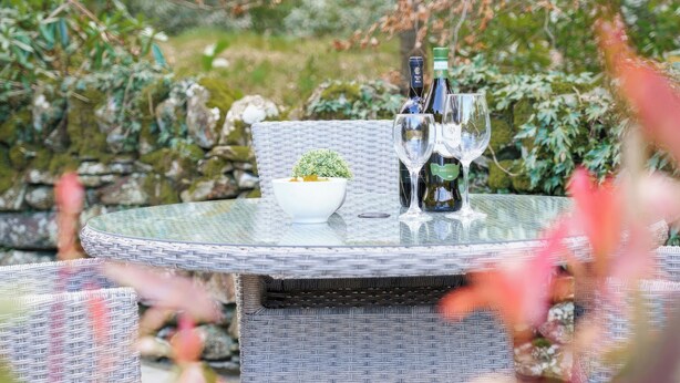 A round glass table with a wicker chair, a small white bowl with a plant, and two wine bottles with glasses.
