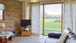 A living room with a leather sofa, a television on a wooden stand, a table with a lamp, and a view of a grassy area through large windows. - SWALLOWS COTTAGE - 24285 (Filey)