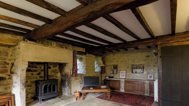 A rustic stone-walled room with a fireplace, wooden beams, stone flooring, a television, and a bookshelf. - 2 bedroom accommodation in Hathersage (Hathersage)