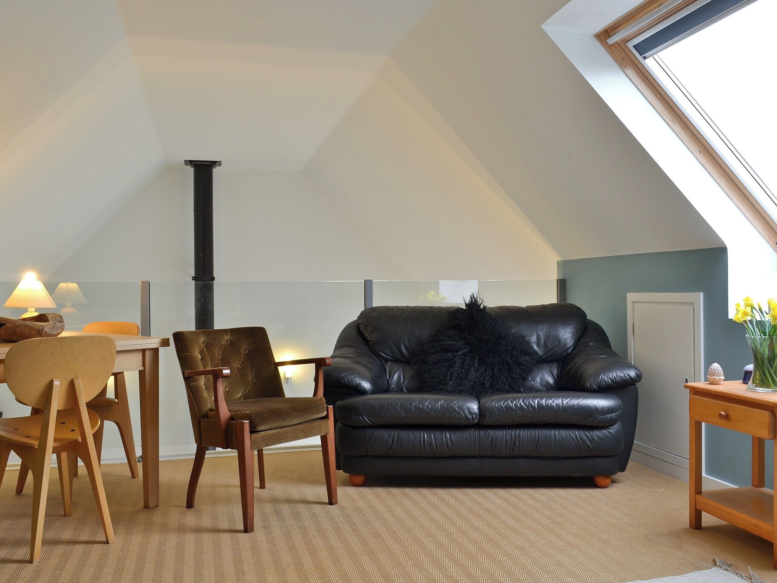 A cozy living room with a black leather sofa, wooden chairs, a small table, and a visible chimney.