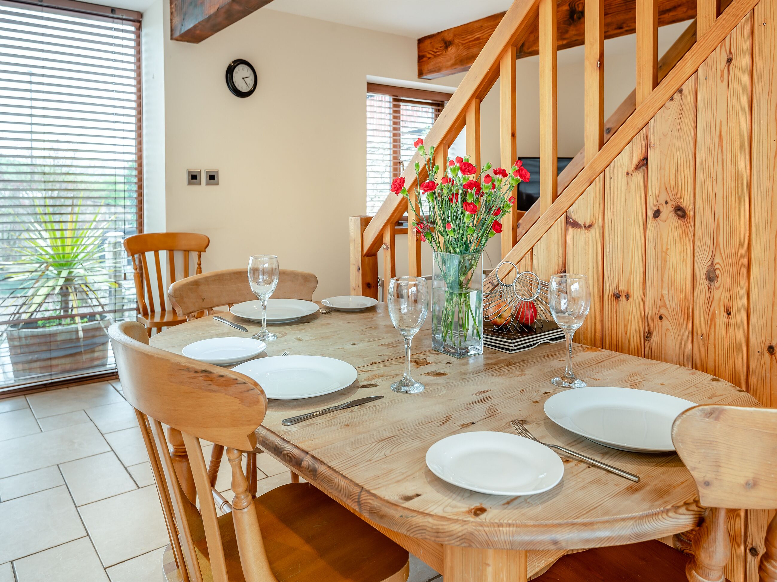 A dining area with a wooden table set for four, surrounded by wooden chairs. A vase with red flowers is on the table, and a clock is on the wall.