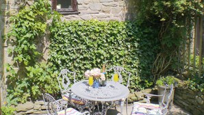 Un patio al aire libre con una mesa metálica redonda y sillas blancas, rodeado de abundante follaje verde.