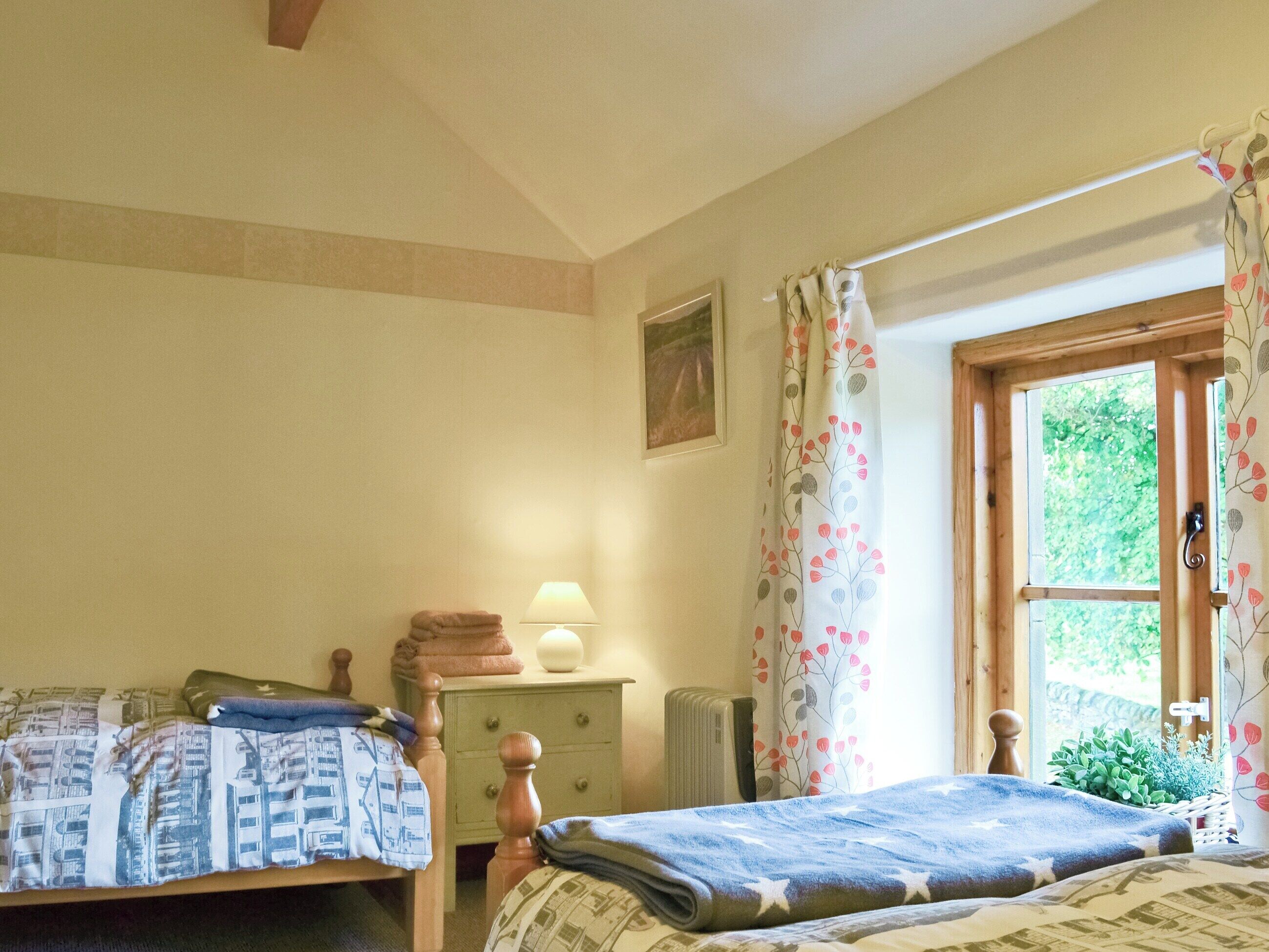 A bedroom with two beds, a nightstand, a lamp, a window with curtains, and a small plant.