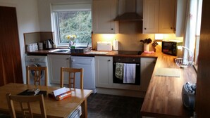 A compact kitchen with a wooden table, chairs, a washing machine, a dishwasher, and a window with a view of greenery. - Tigh Na Fraoch, (Skeabost)