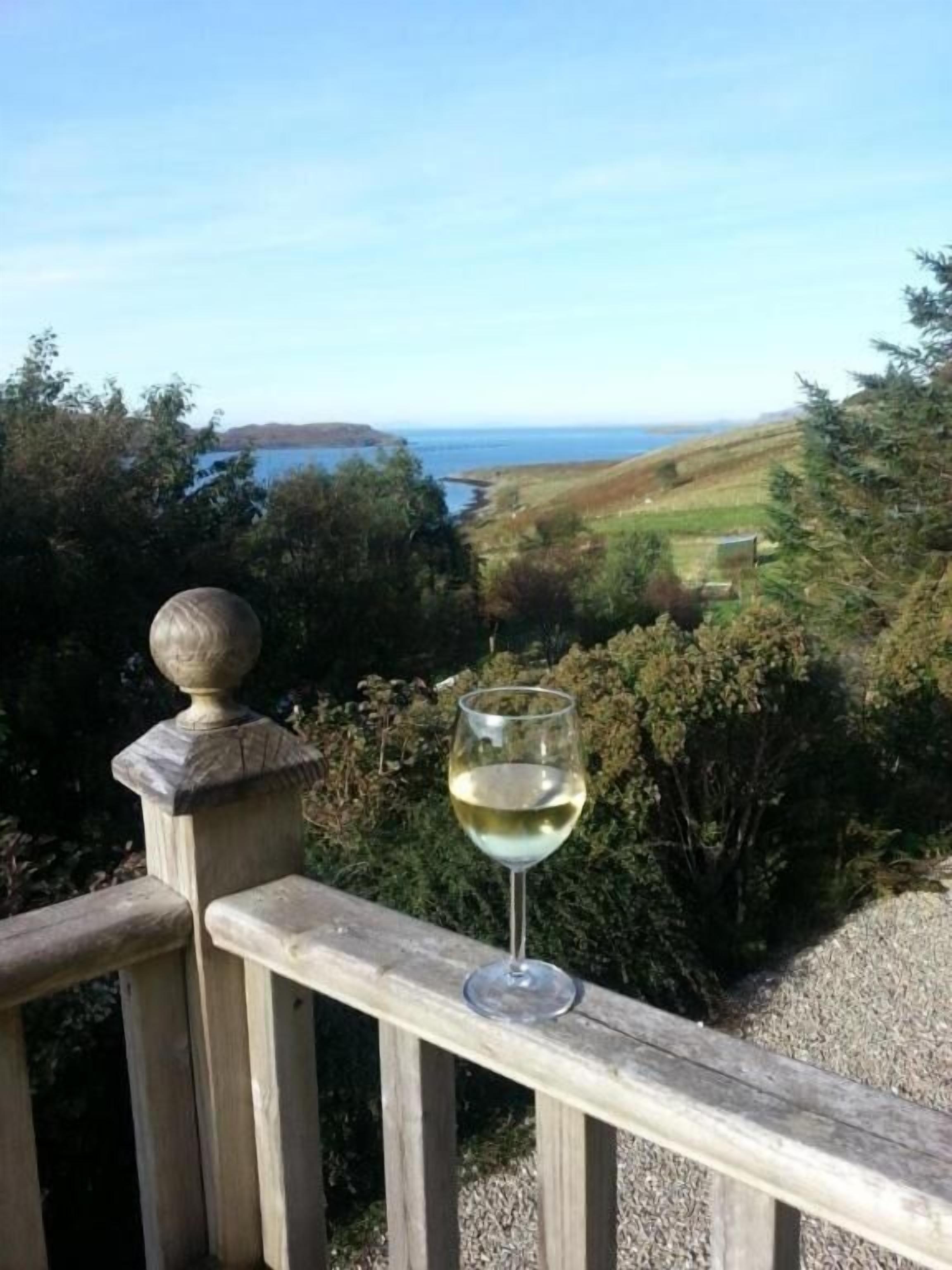 A glass of white wine on a wooden railing overlooking a coastal landscape with hills and a body of water.