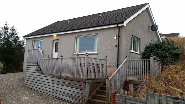 A single-story house with a sloped roof, a front porch with a wooden railing, and a set of wooden stairs leading to the entrance. - Tigh Na Fraoch, (Skeabost)