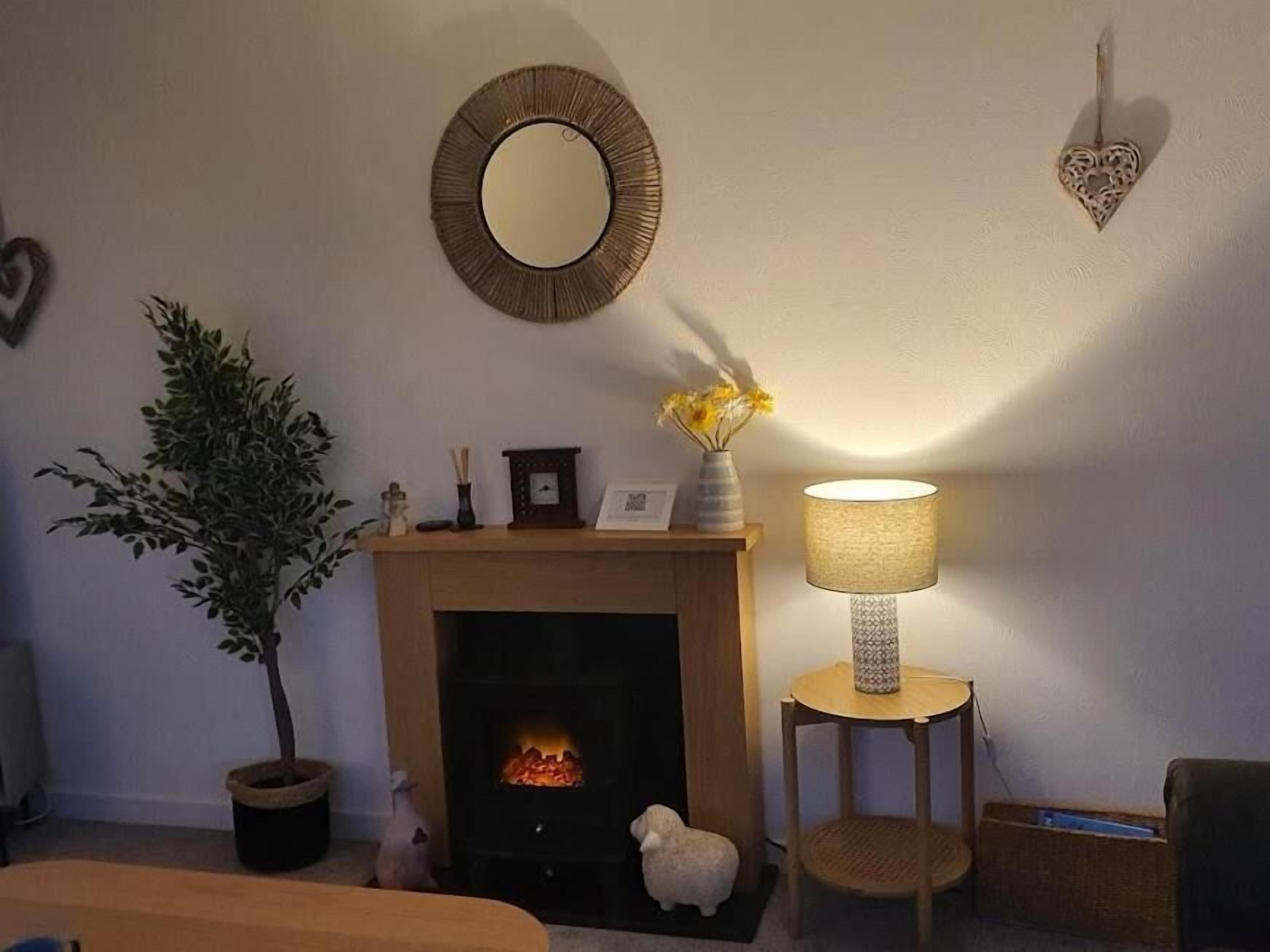 A cozy living room with a fireplace, a round mirror, a potted plant, a lamp, and a heart-shaped decoration on the wall.