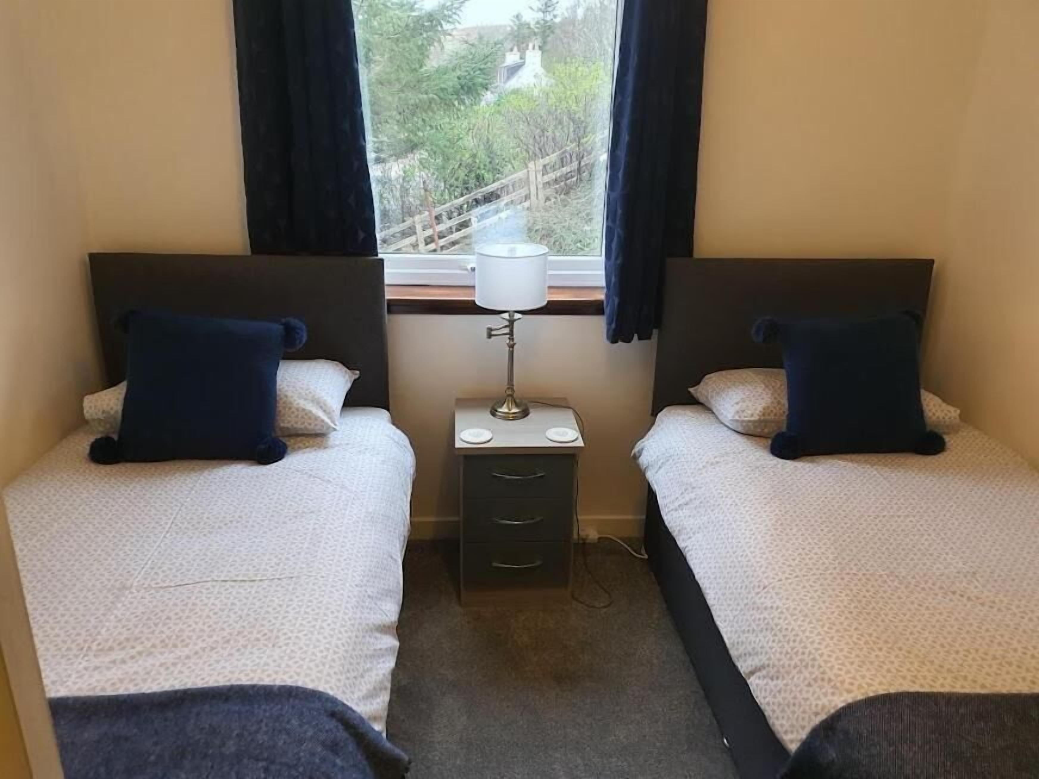 A twin room with two beds, a bedside table with a lamp, and a window with curtains.