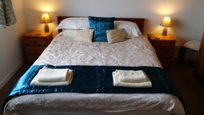 A neatly made bed with a patterned bedspread, two pillows, and two folded towels. There are two bedside tables with lamps, and a wooden headboard. - Tigh Na Fraoch, (Skeabost)