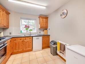A kitchen with wooden cabinets, a dishwasher, a trash bin, a window with plants, and a clock on the wall.