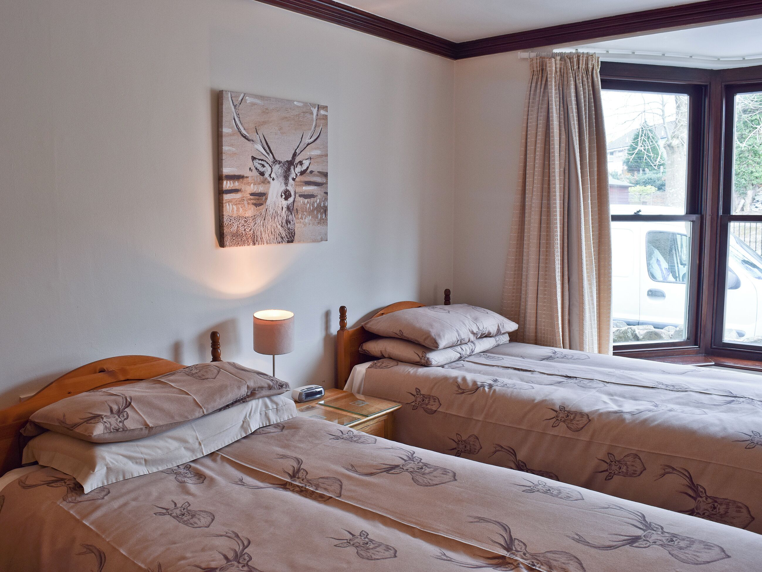 A bedroom with two beds, a bedside table with a lamp, a painting on the wall, and a window with curtains.
