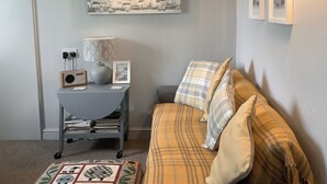 A cozy living room with a grey sofa, patterned cushions, a small side table with a radio, a vase, and a framed picture.