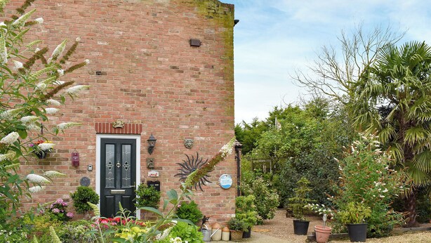 A brick building with a black door, surrounded by a garden with various plants and flowers.