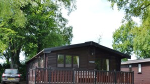 Une cabane en bois avec une terrasse entourée d’une clôture, une voiture garée devant et une pancarte indiquant « Glenmore ».