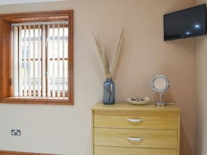 A room with a wooden dresser, a vase with dried plants, a bowl, a round mirror, and a TV mounted on the wall.